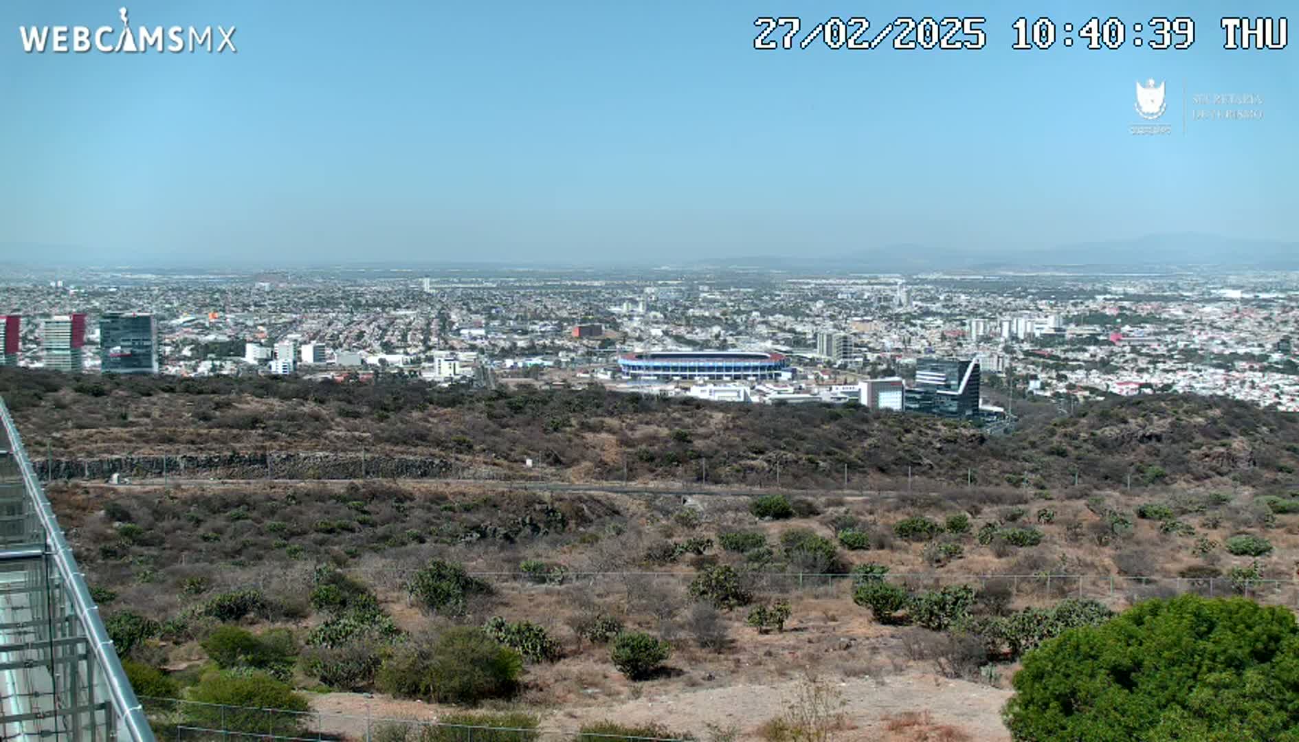 A sprawling city is visible in the distance under a clear blue sky, viewed from a high vantage point overlooking a dry, scrubby hillside.