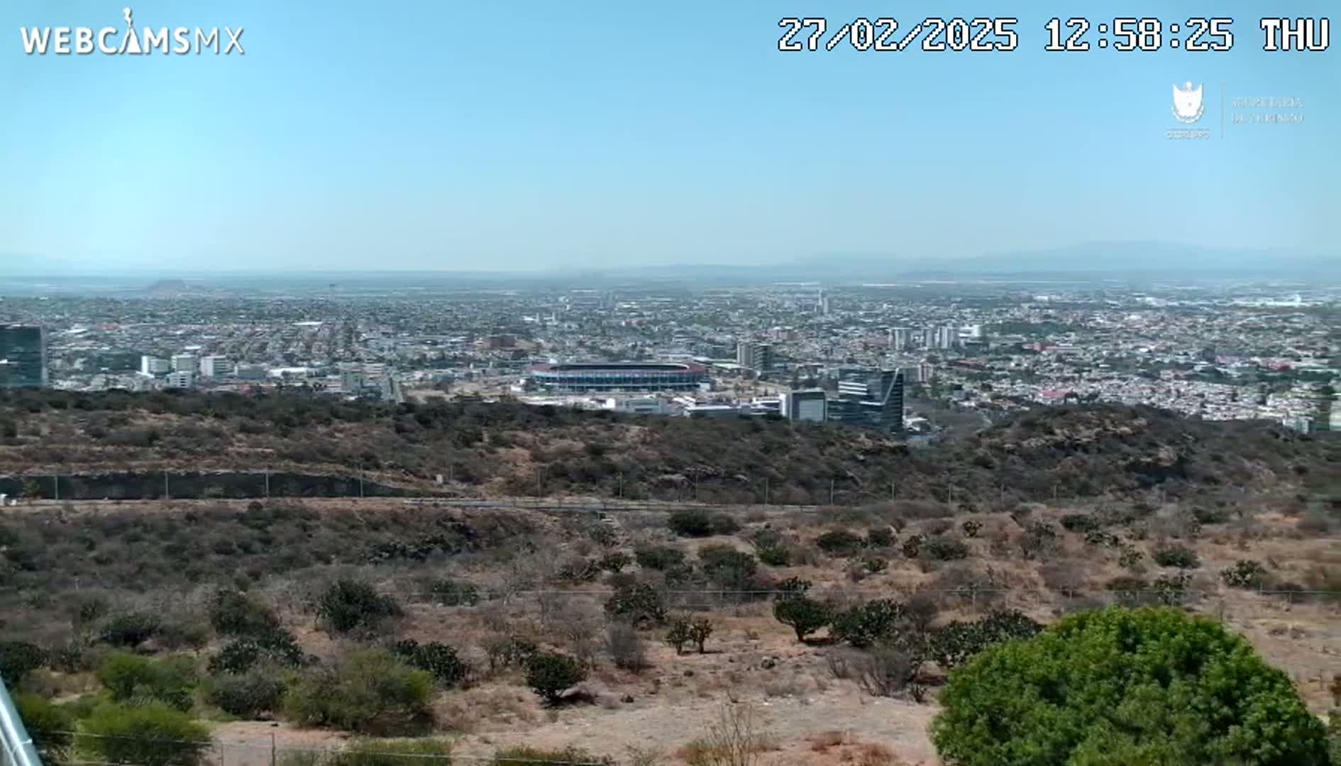 A high-angle, long shot on a clear day shows a sprawling city with a stadium visible, surrounded by scrubby, dry hills.