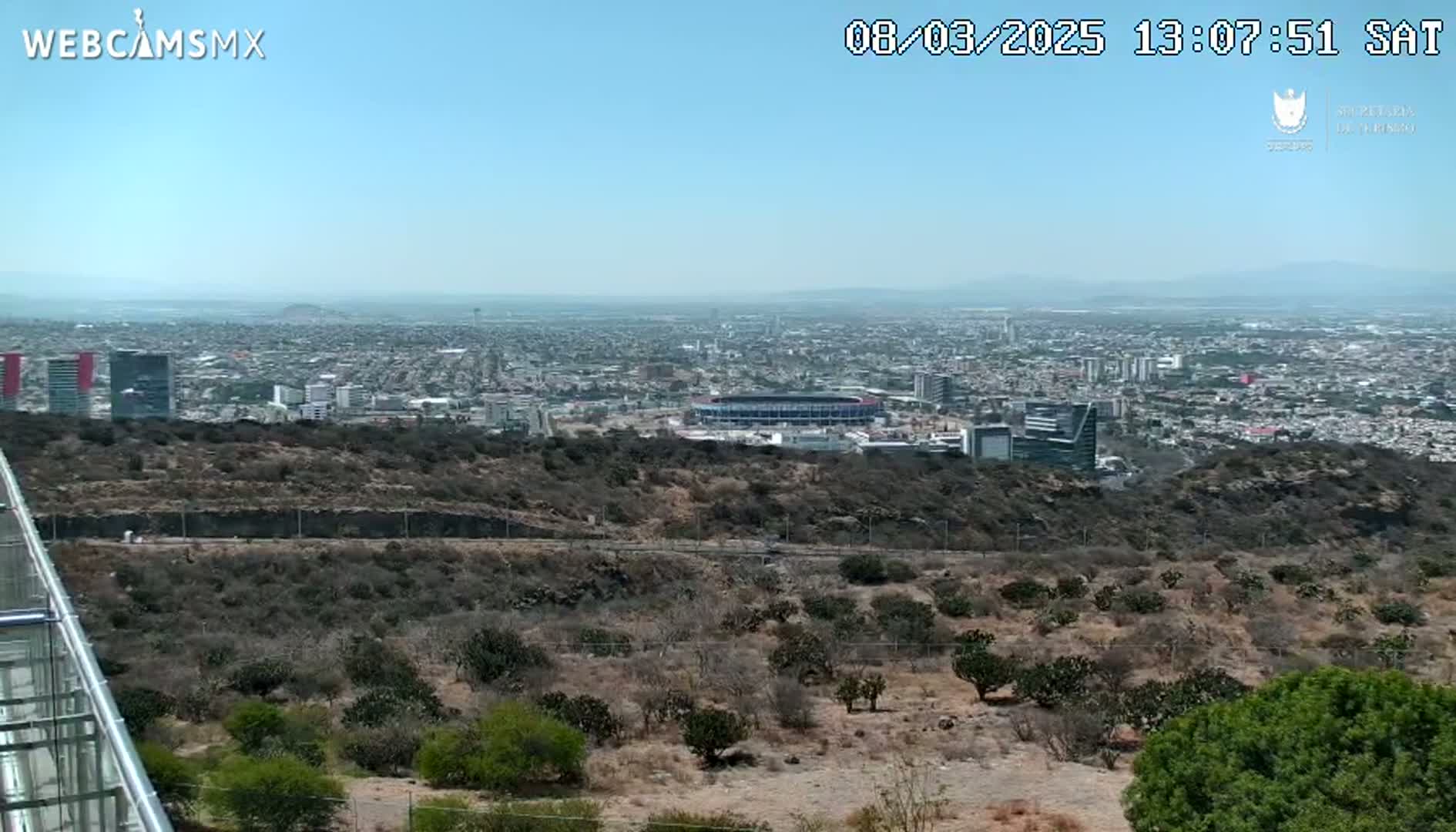 A high-angle, long shot on a clear day shows a large city sprawling across a flat plain, partially obscured by a rocky, scrub-covered hillside in the foreground, with a stadium visible in the mid-ground.