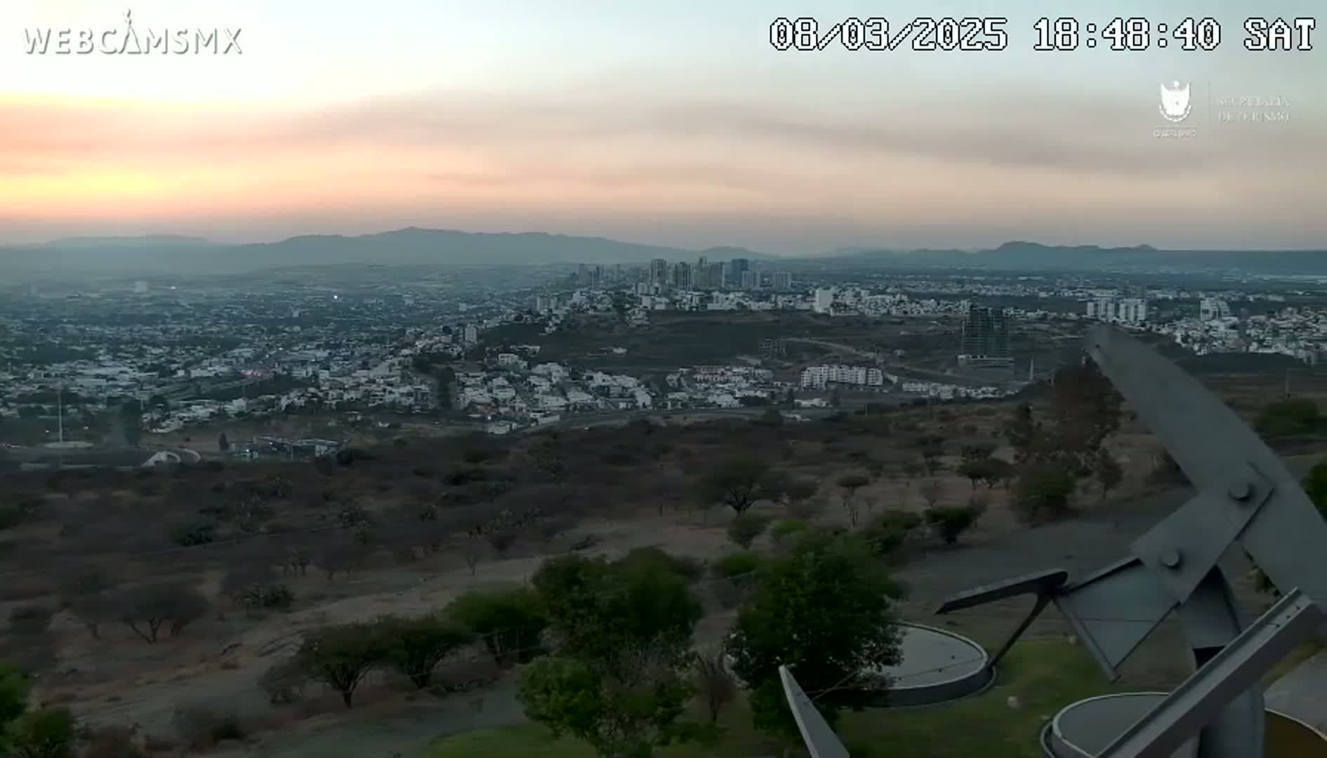 A sprawling city at sunset is viewed from a high vantage point, with a large metal sculpture in the foreground.