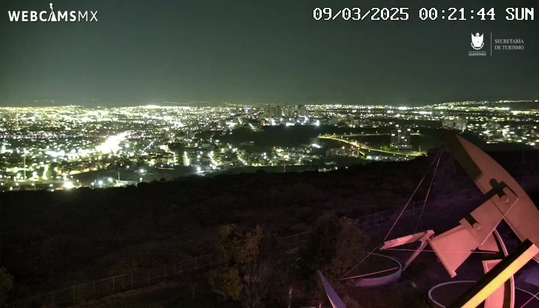 A nighttime view from a hilltop shows a sprawling city illuminated below, under a clear sky.
