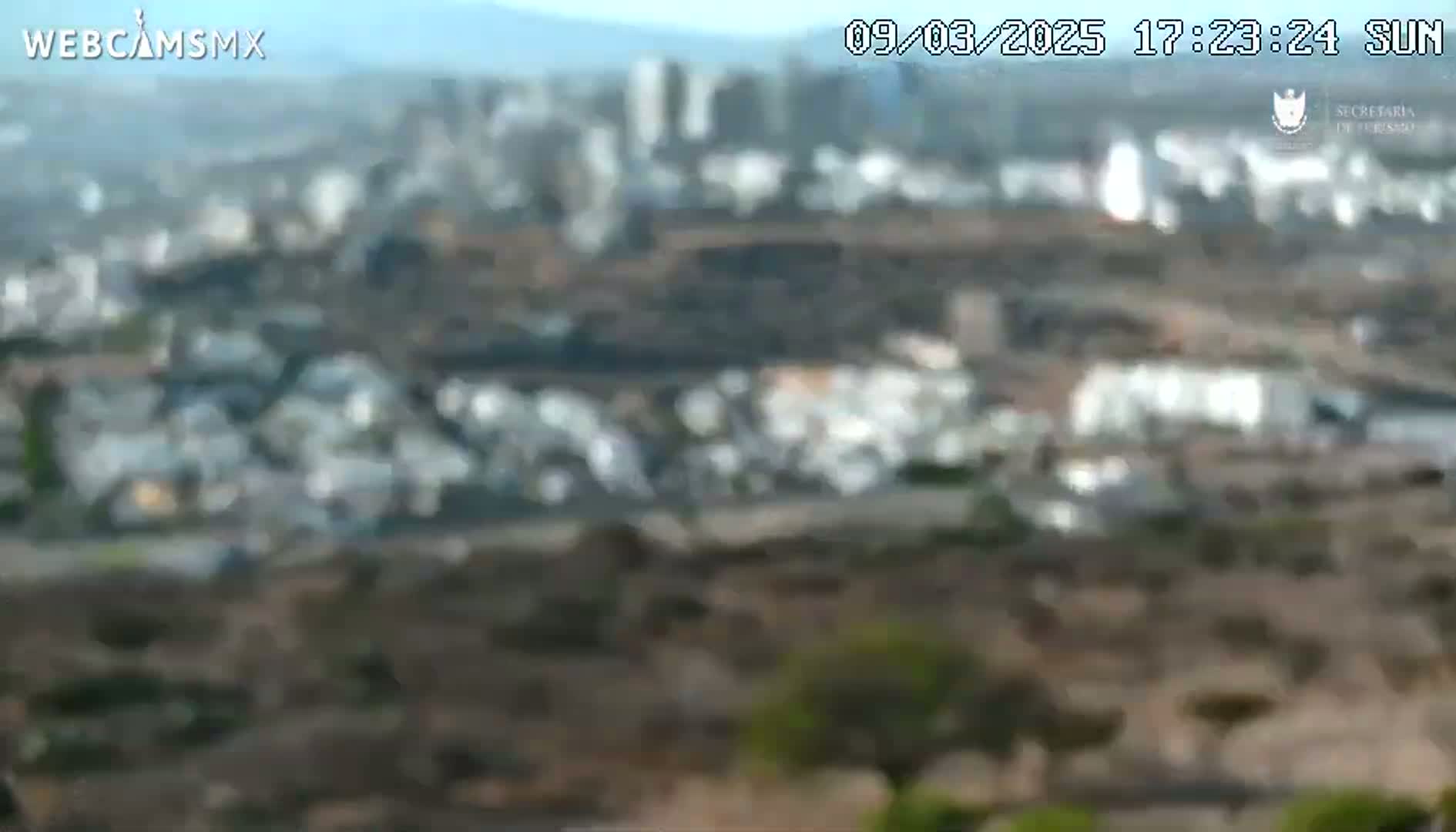A blurry, long shot of a city in the distance under a mostly clear sky, viewed from a high vantage point overlooking a dry, hilly landscape.