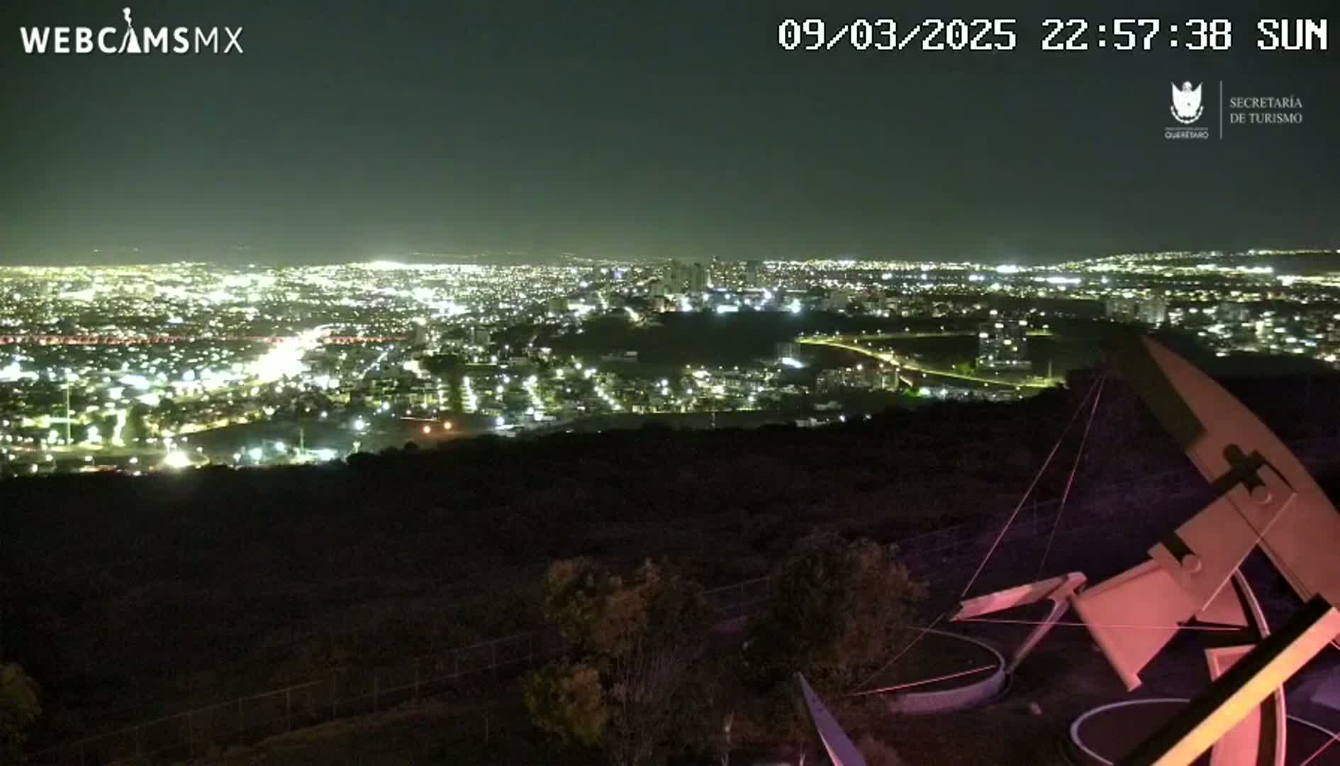 A nighttime view from a hilltop shows a sprawling city illuminated below, with a large, abstract metal sculpture in the foreground under a clear sky.