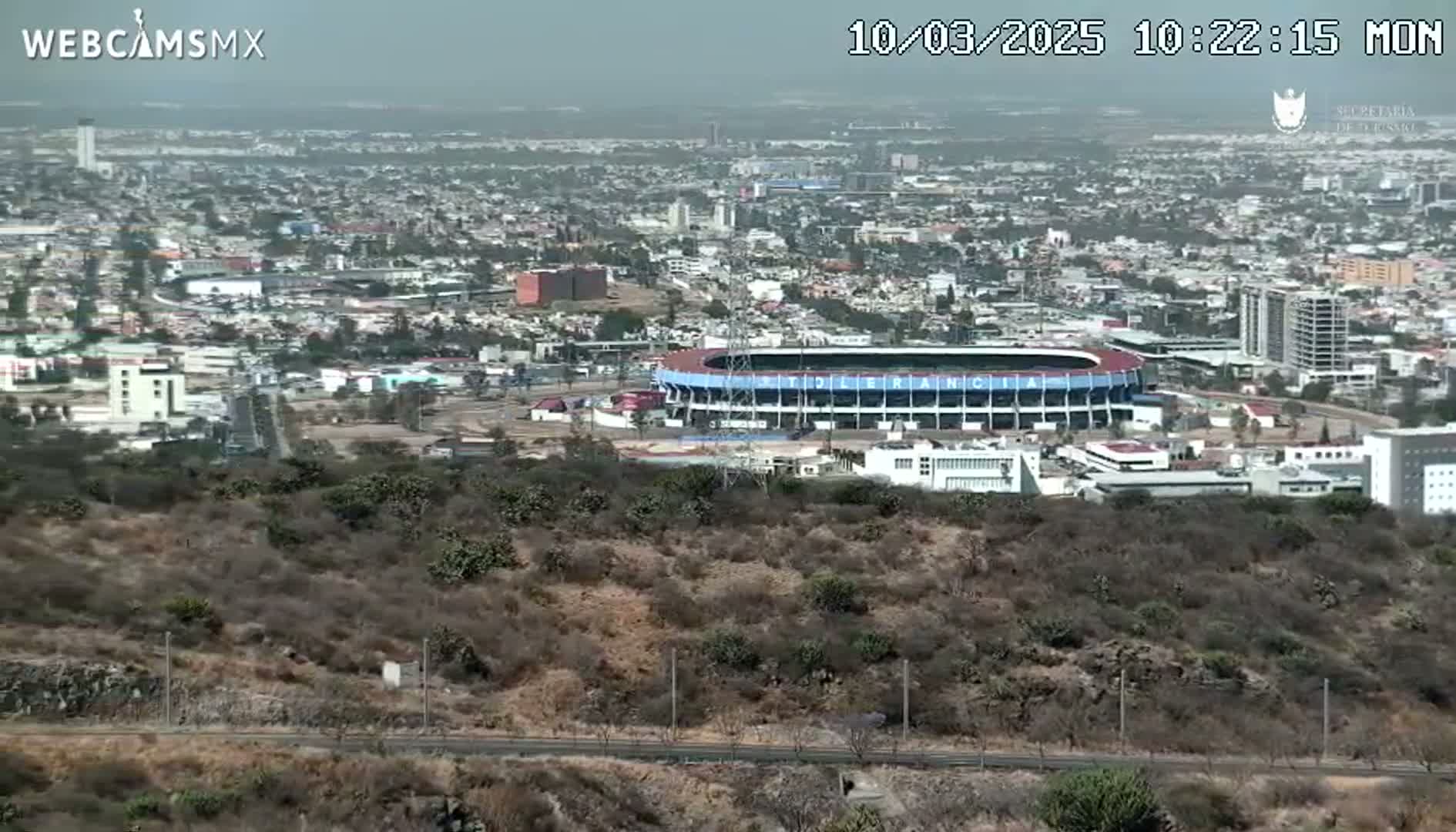 A sprawling city, partially obscured by hazy air, features a large stadium nestled amongst low-lying scrubland.
