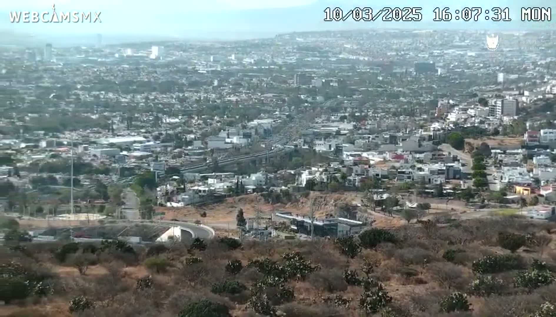 A hazy, high-angle view shows a sprawling city extending to distant mountains under a mostly clear sky.