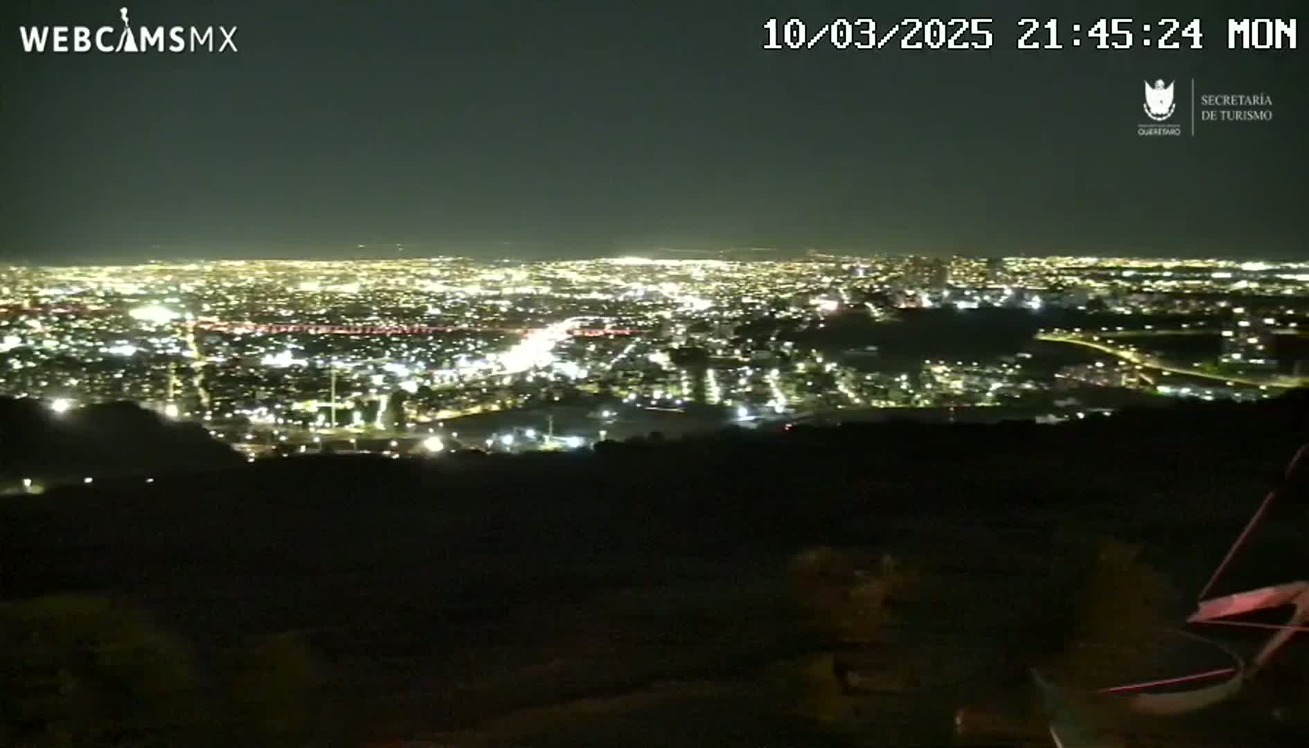 A nighttime, high-angle view shows a sprawling city illuminated by countless lights, observed from a dark, hilly vantage point under a clear sky.