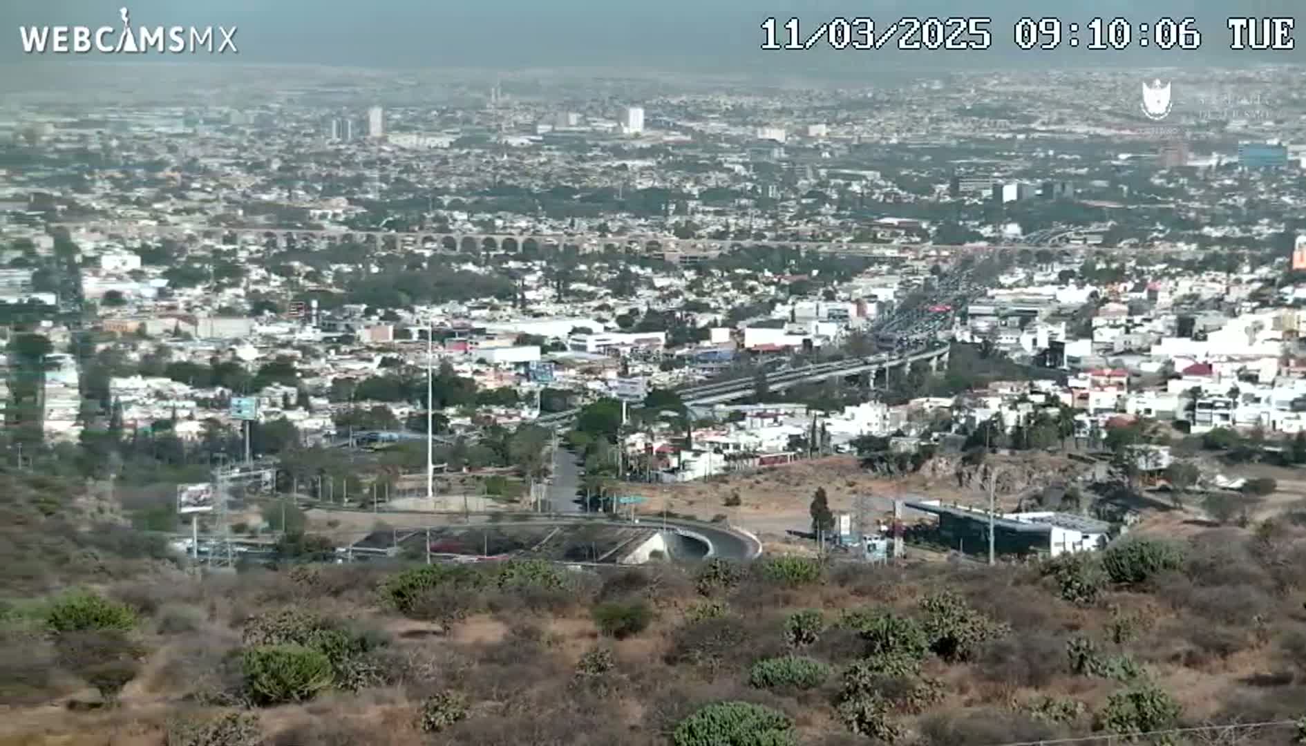 A sprawling city view on a hazy day is seen from a hilltop, revealing a network of roads, buildings, and a large highway overpass.
