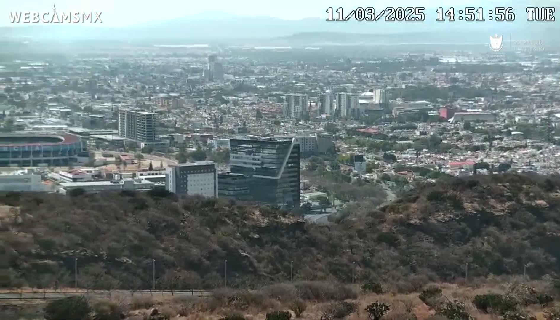 A hazy view from a hilltop shows a sprawling city with a stadium, numerous buildings, and dry, scrub-covered hills in the foreground.