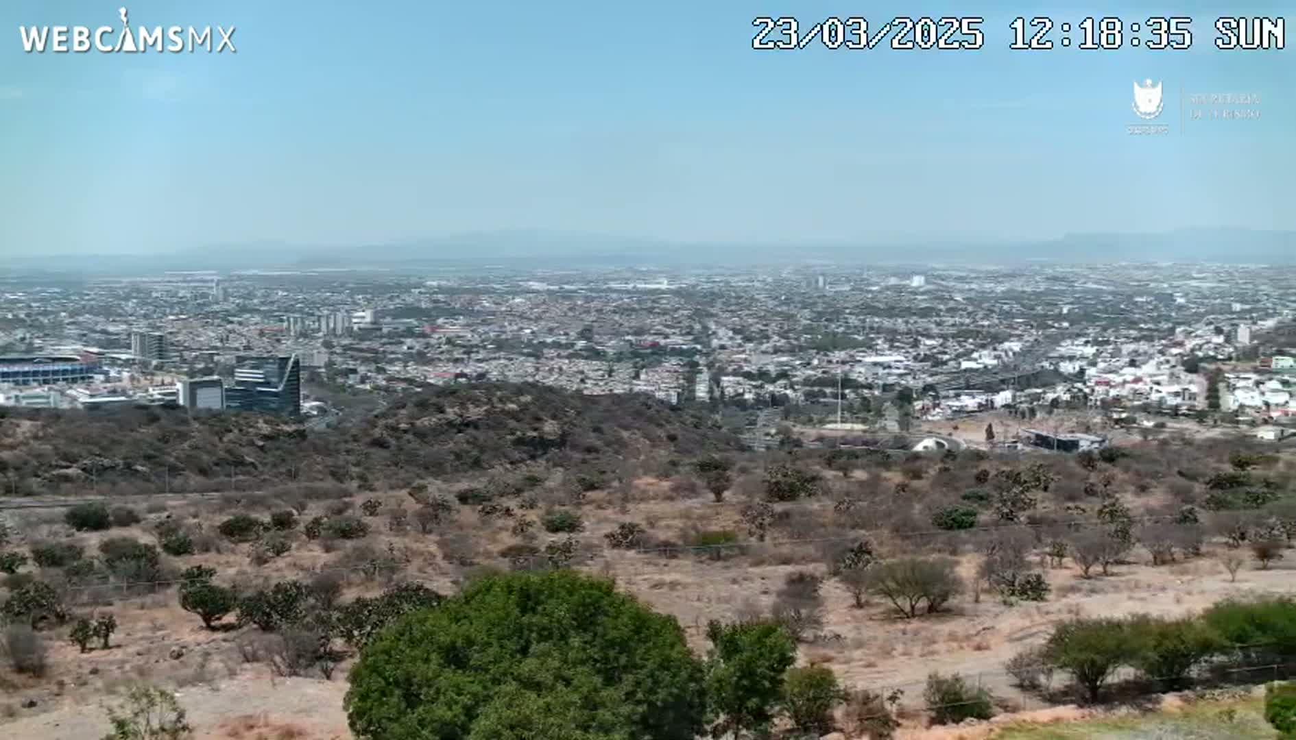 A sprawling city is visible in the distance under a clear, sunny sky, viewed from a high vantage point overlooking scrubby, dry hills.