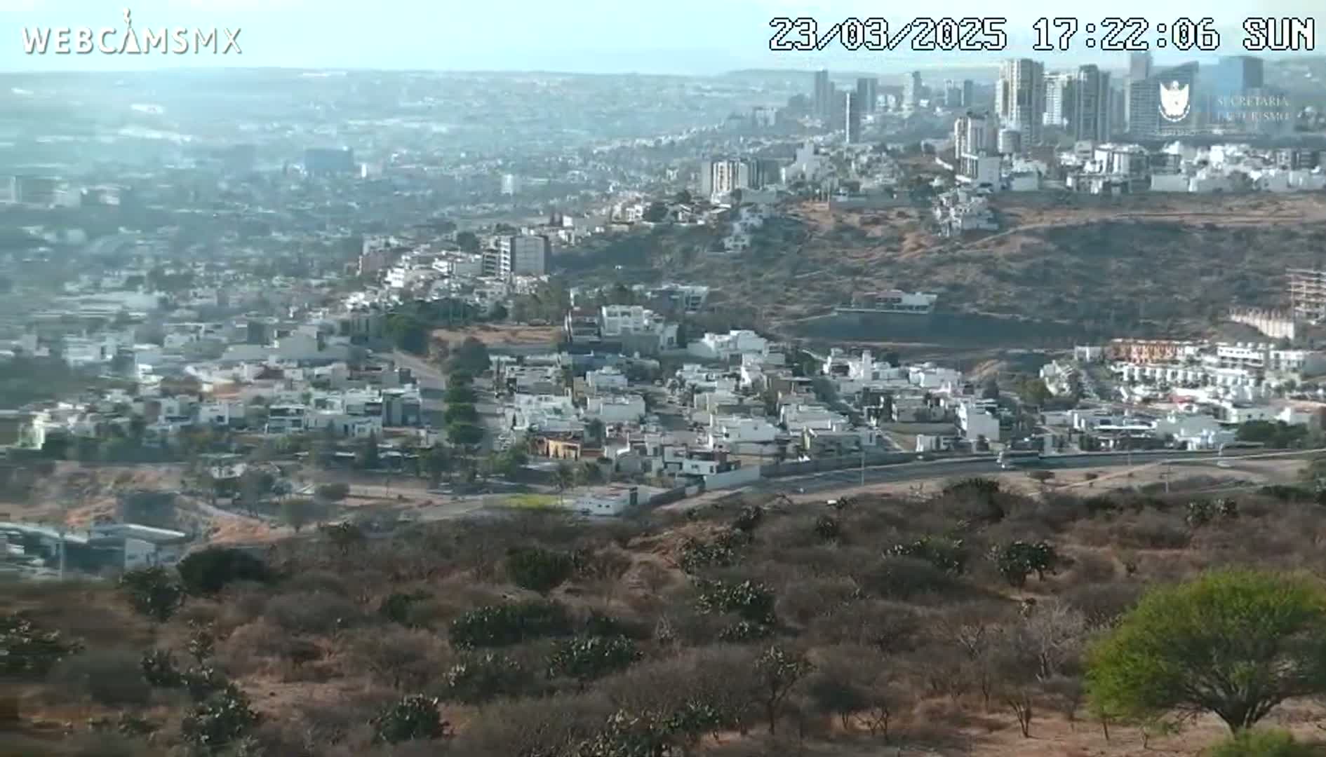 A sprawling city with white buildings on a hillside is visible under a hazy, sunny sky, observed from an elevated vantage point overlooking scrubland.