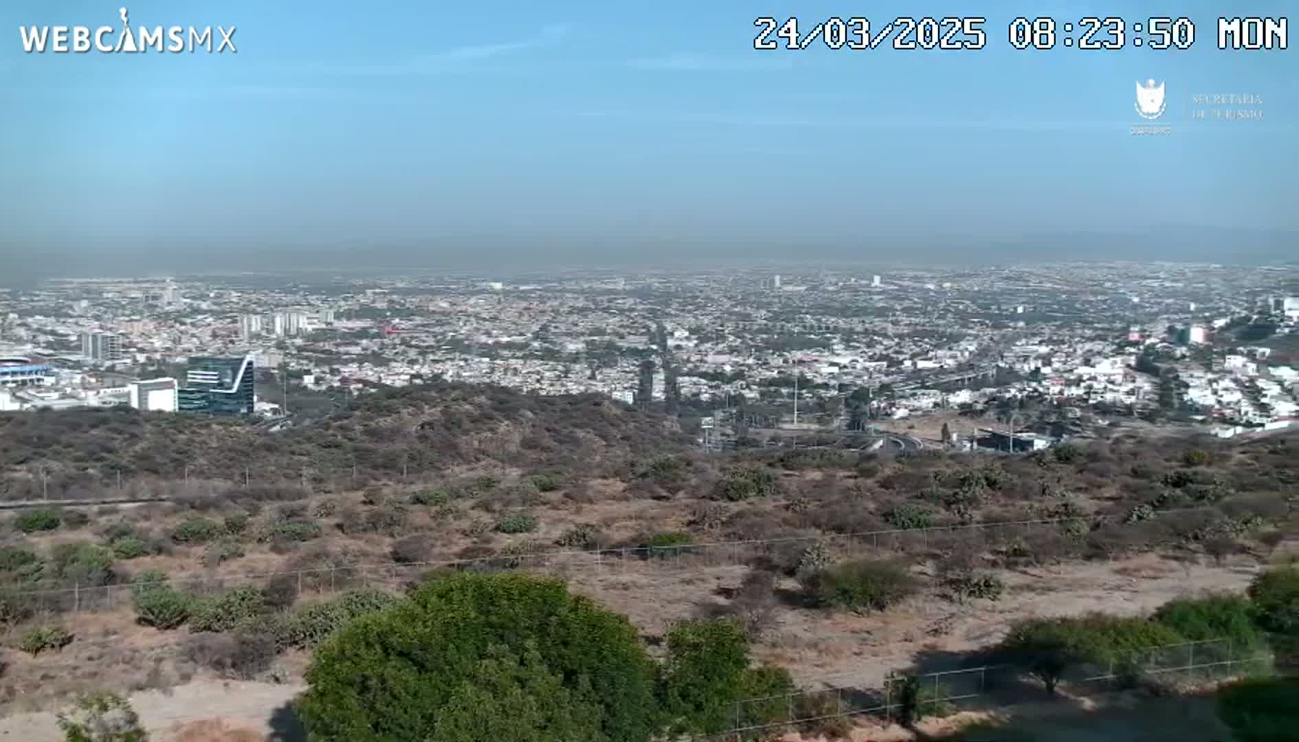 A sprawling city is visible in the distance under a clear, hazy sky, viewed from a high vantage point overlooking a scrubby, hilly landscape.