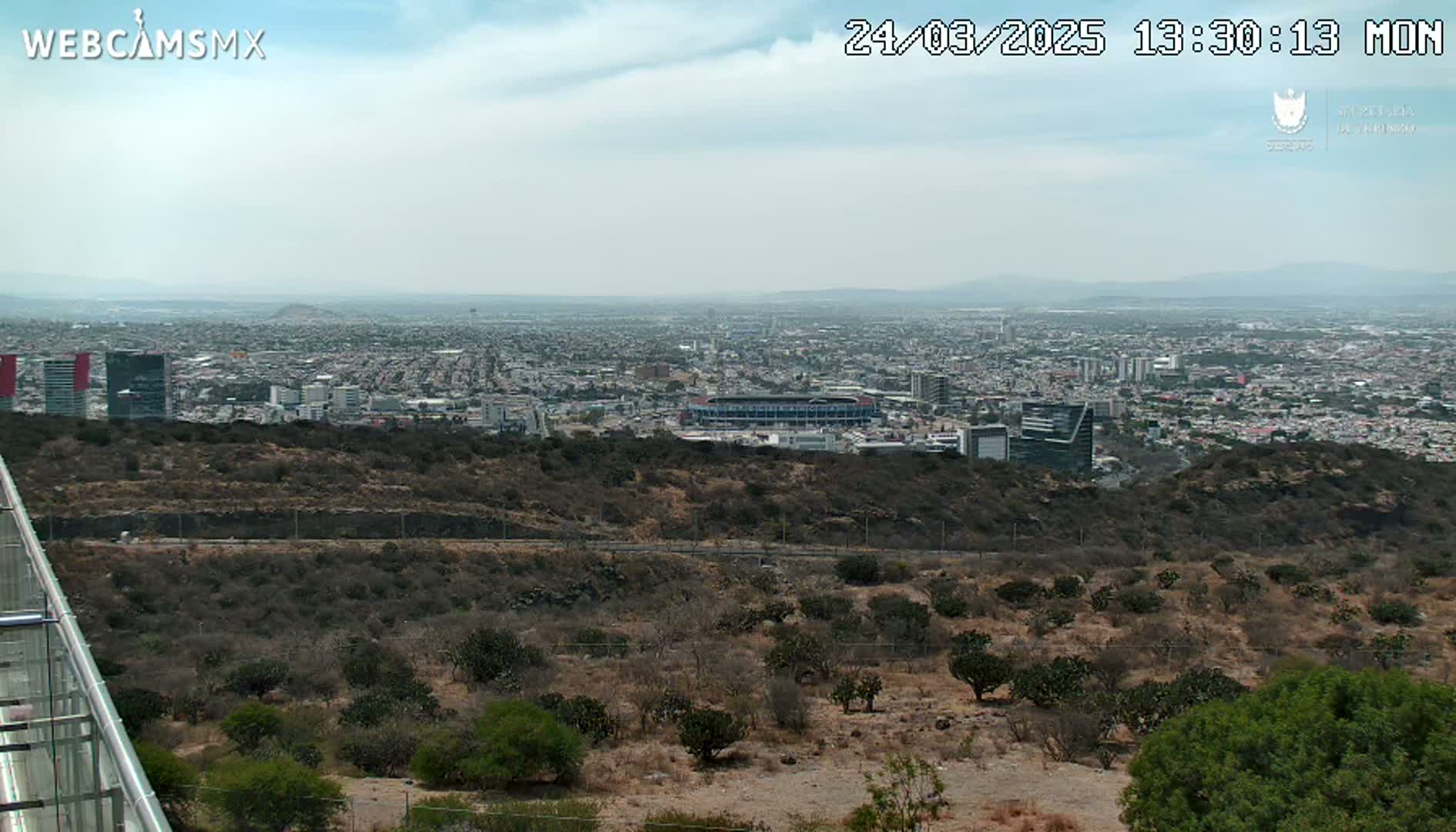 A sprawling city is visible in the distance under a mostly cloudy sky, viewed from a higher vantage point overlooking scrubby, dry hills and a glass railing.