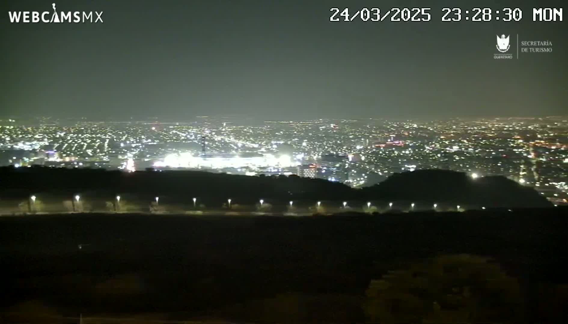 A nighttime, hazy view from a hilltop overlooking a brightly lit city.