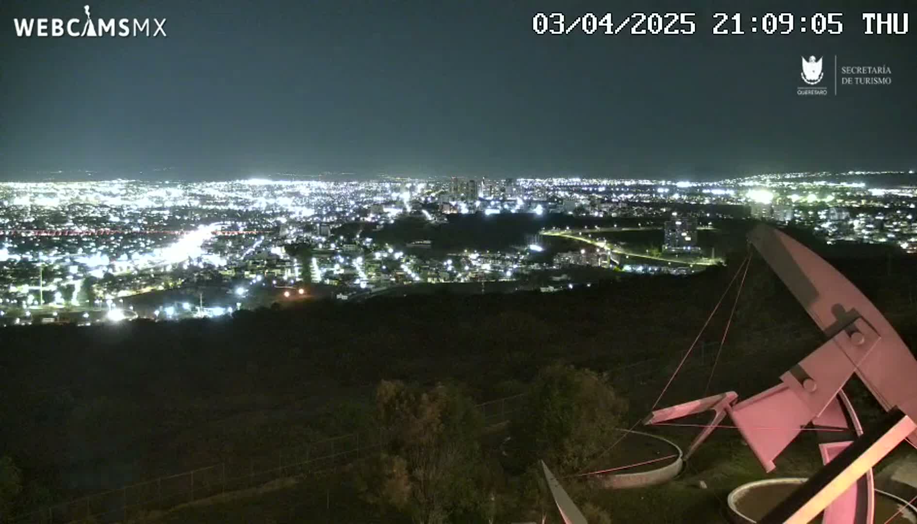 A nighttime view from a hilltop shows a sprawling city illuminated below, with a large metal sculpture in the foreground under a clear, dark sky.