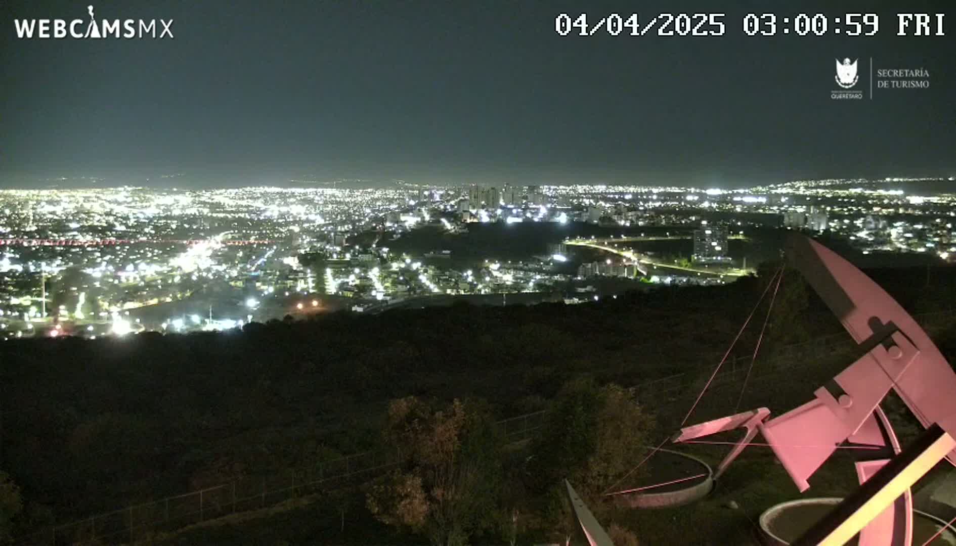 A nighttime view from an elevated position shows a sprawling city illuminated below, with a large, pinkish-white metal structure in the foreground under a clear, dark sky.
