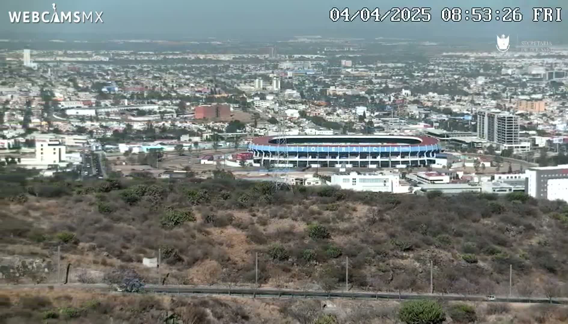 A sprawling city, under a hazy sky, is viewed from a hilltop, showcasing a large stadium in the foreground.