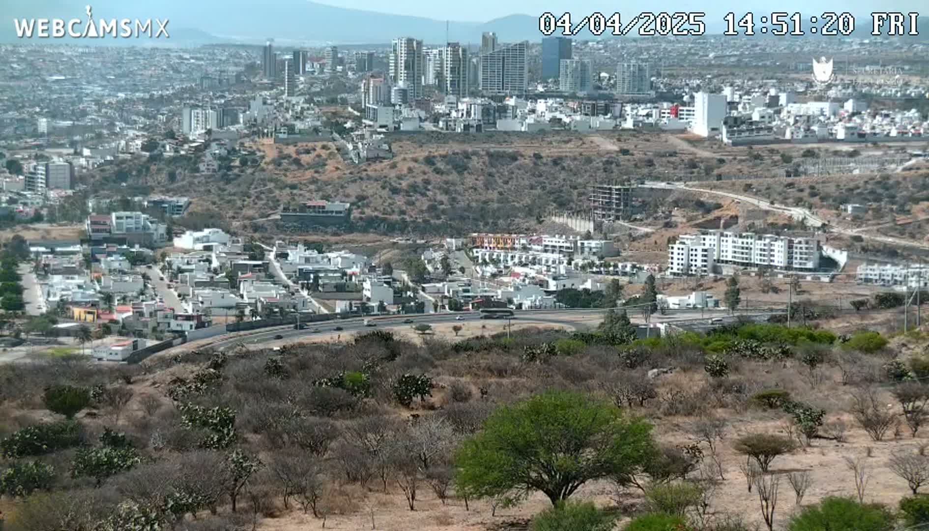 A high-angle, long shot shows a sprawling city on a sunny day, with a road curving through a dry, scrubby landscape in the foreground.
