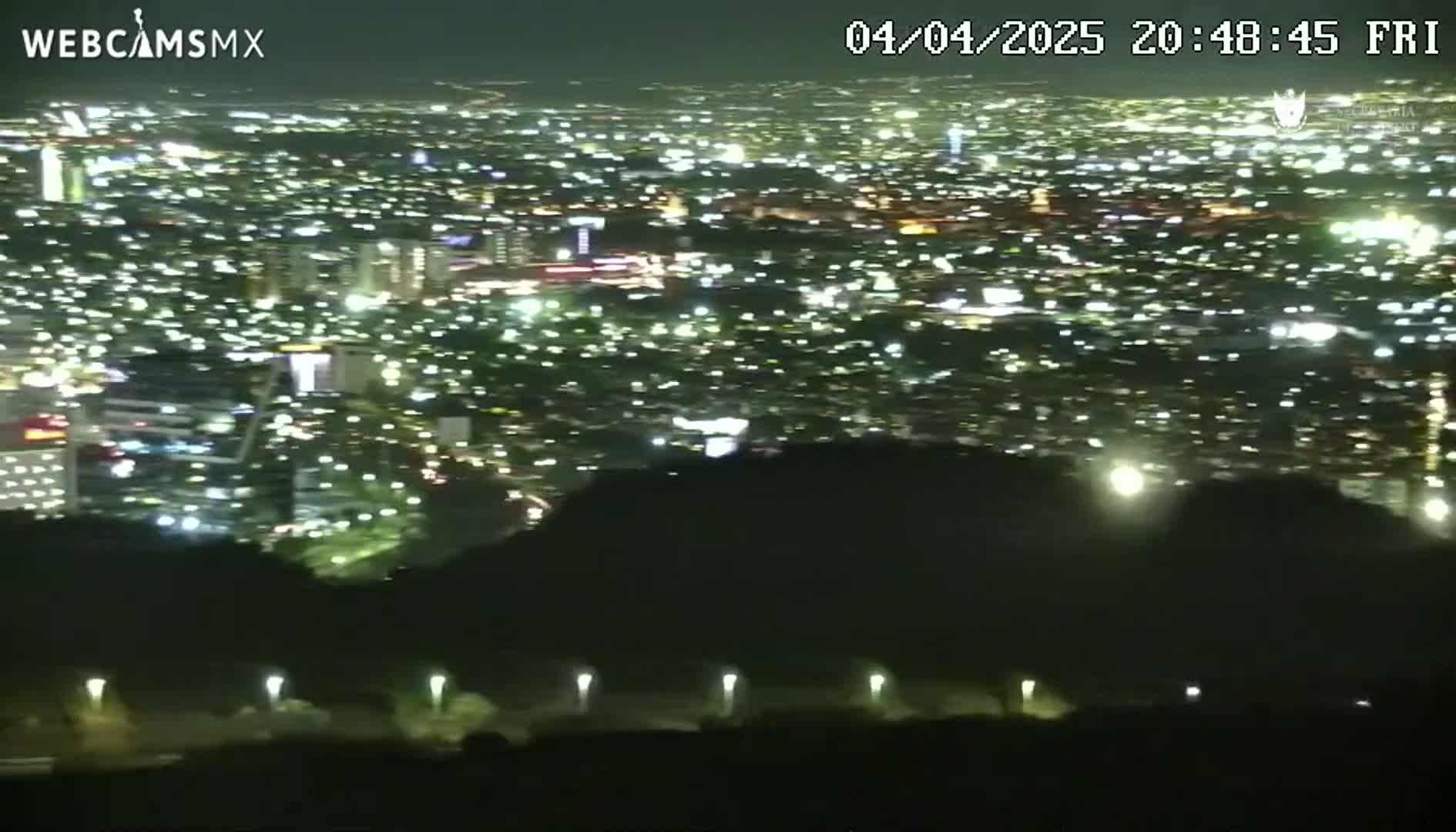 A nighttime view from an elevated position shows a sprawling city illuminated by countless lights, with dark hills or mountains in the foreground.