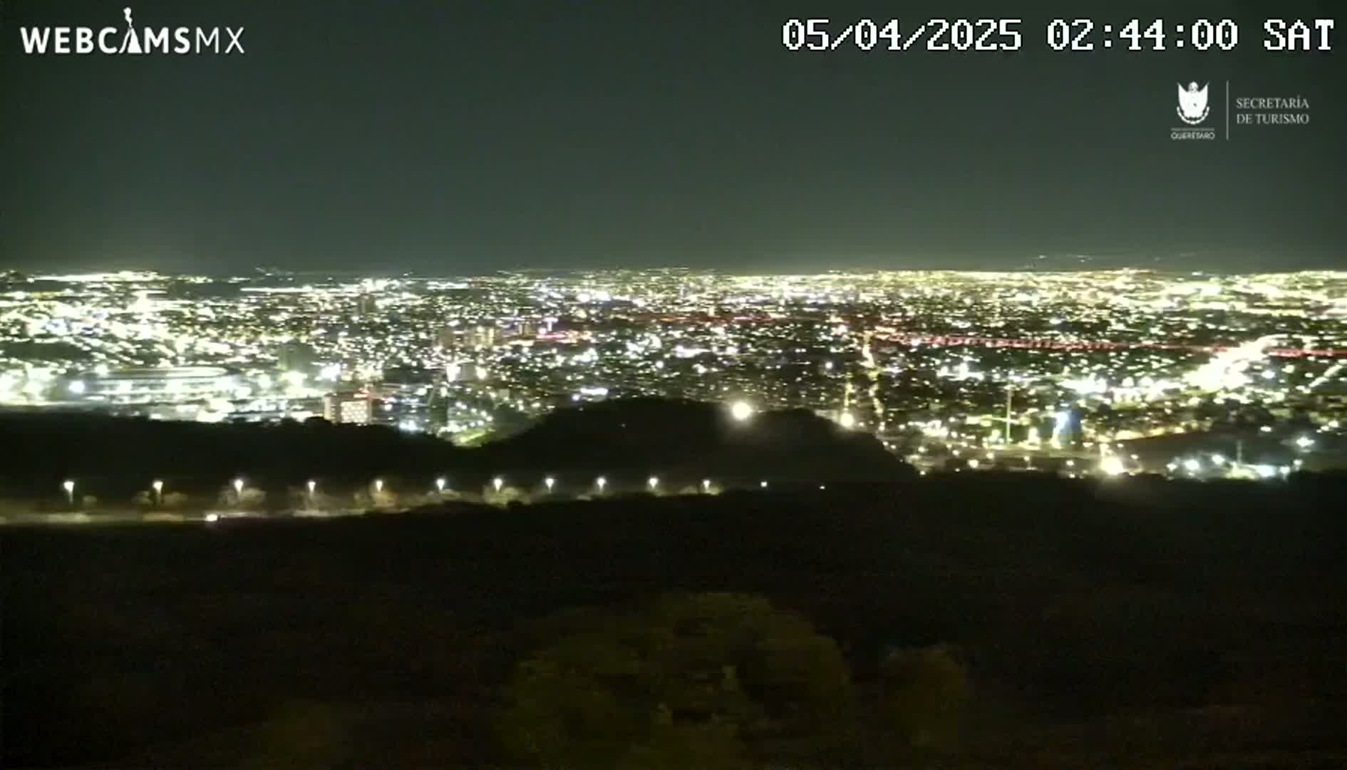 A nighttime view from an elevated vantage point shows a sprawling city illuminated by numerous lights, with a dark, hilly foreground.