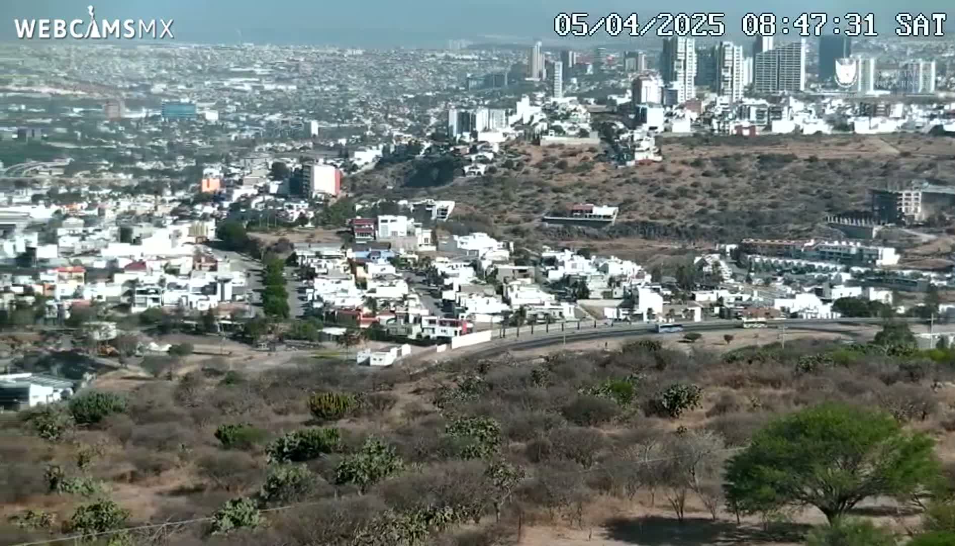 An elevated view on a clear day shows a sprawling city with low-rise buildings and a hilly, sparsely vegetated landscape in the foreground.