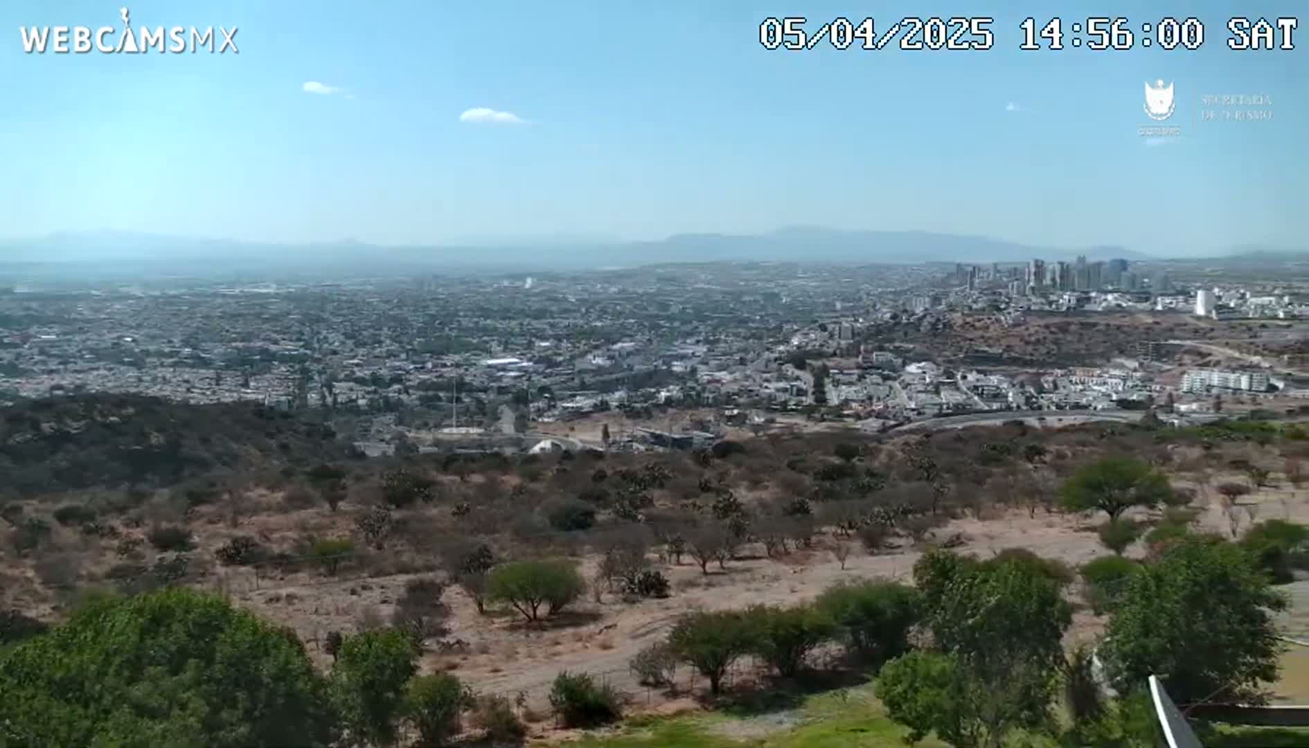 A sprawling city is visible in the distance under a clear, sunny sky, viewed from a high vantage point overlooking arid, scrubby hills and vegetation in the foreground.