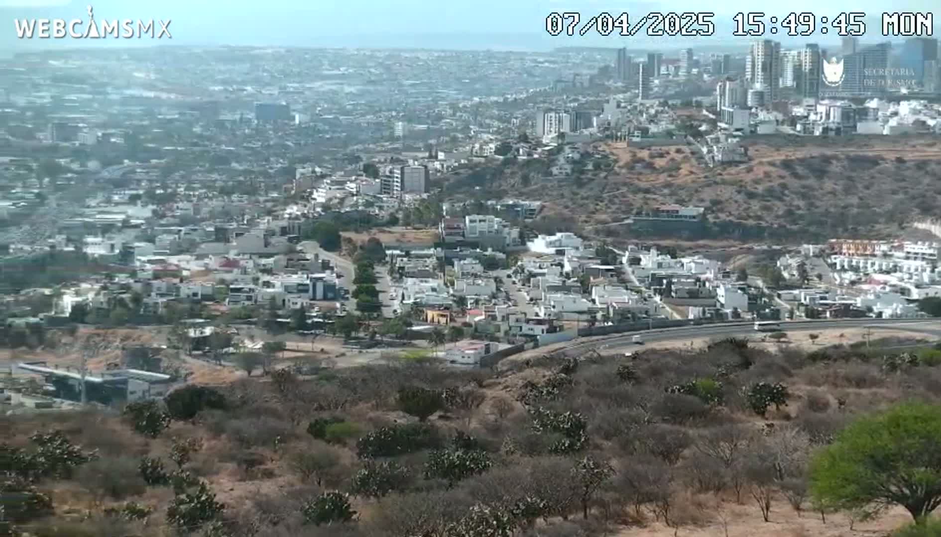 A high-angle, long shot on a sunny day shows a sprawling city extending to the horizon, with a foreground of dry, scrubby vegetation and a curving road.