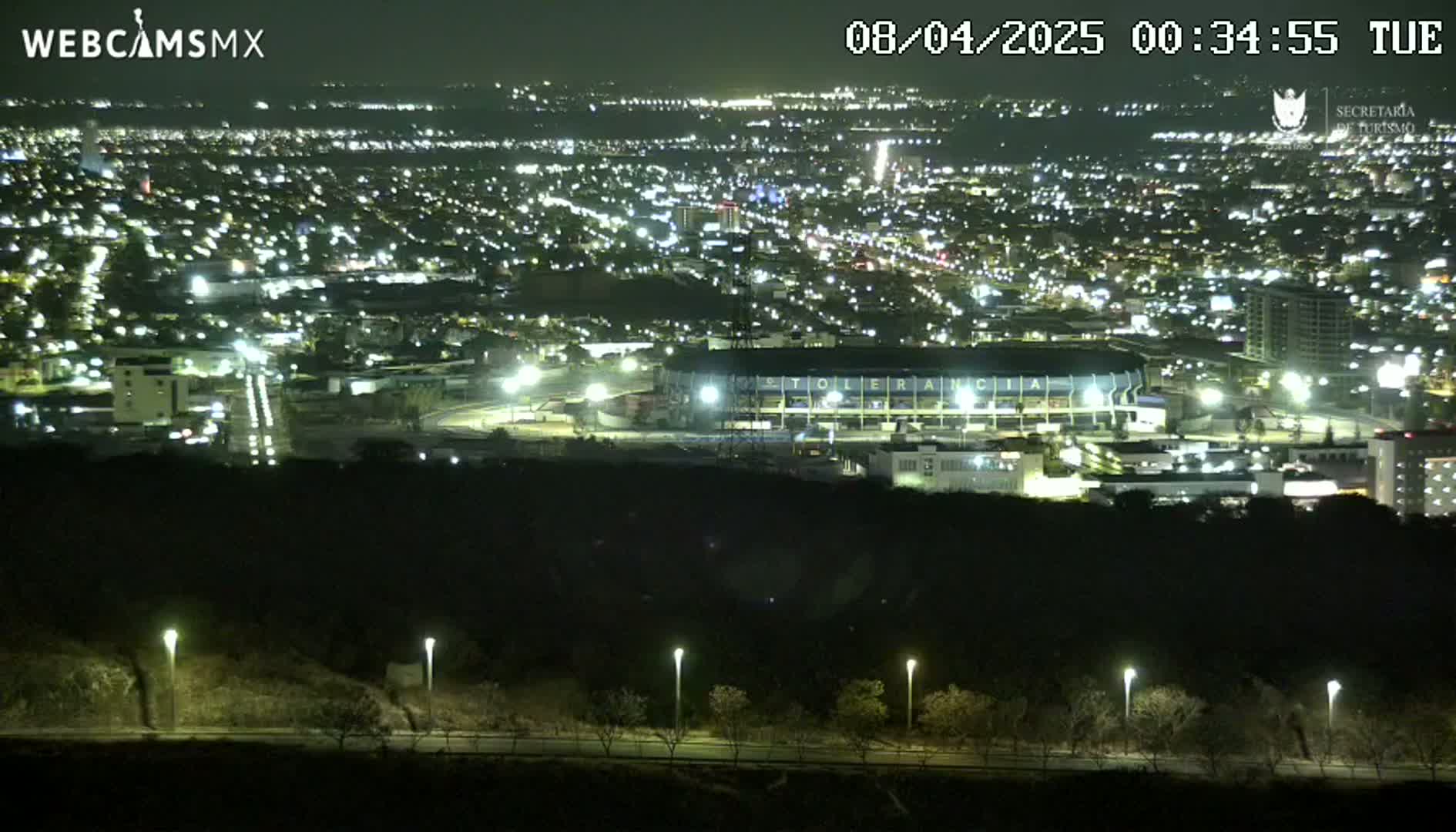 A nighttime, high-angle view shows a brightly lit city sprawling across a valley, including a large stadium with lights on, and a dark hill in the foreground.