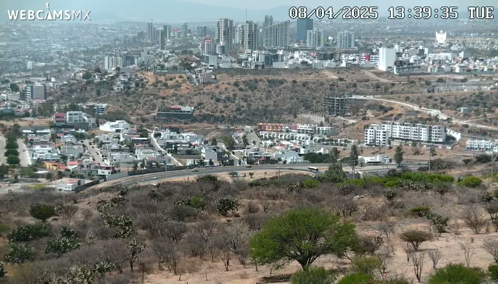 A sprawling city with numerous high-rise buildings is visible in the distance under a hazy, sunny sky, overlooking a lower area of residential homes and scrub brush.
