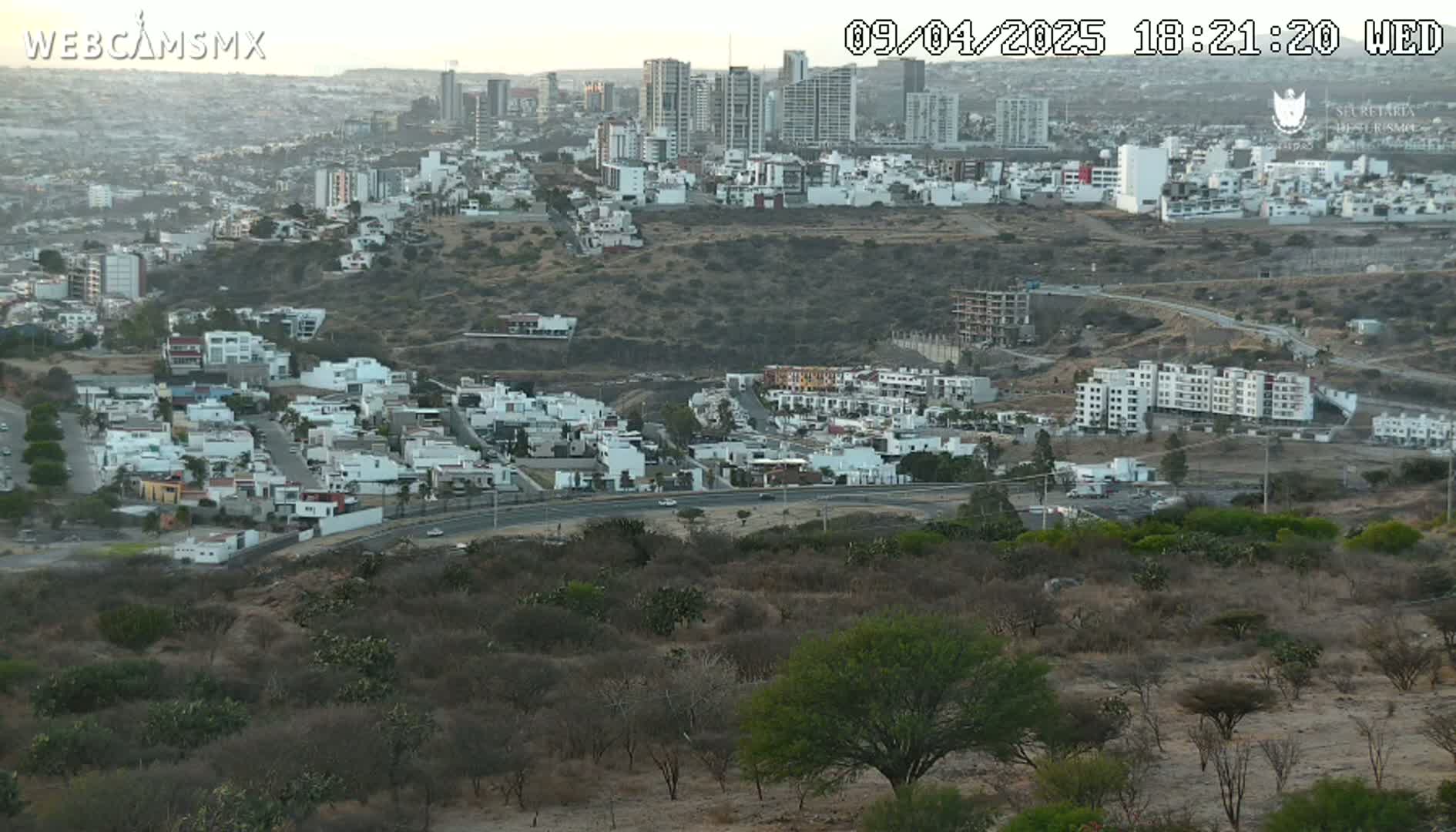 An elevated view of a city with light-colored buildings, a highway, and sparse, dry-looking vegetation under a dusky sky.