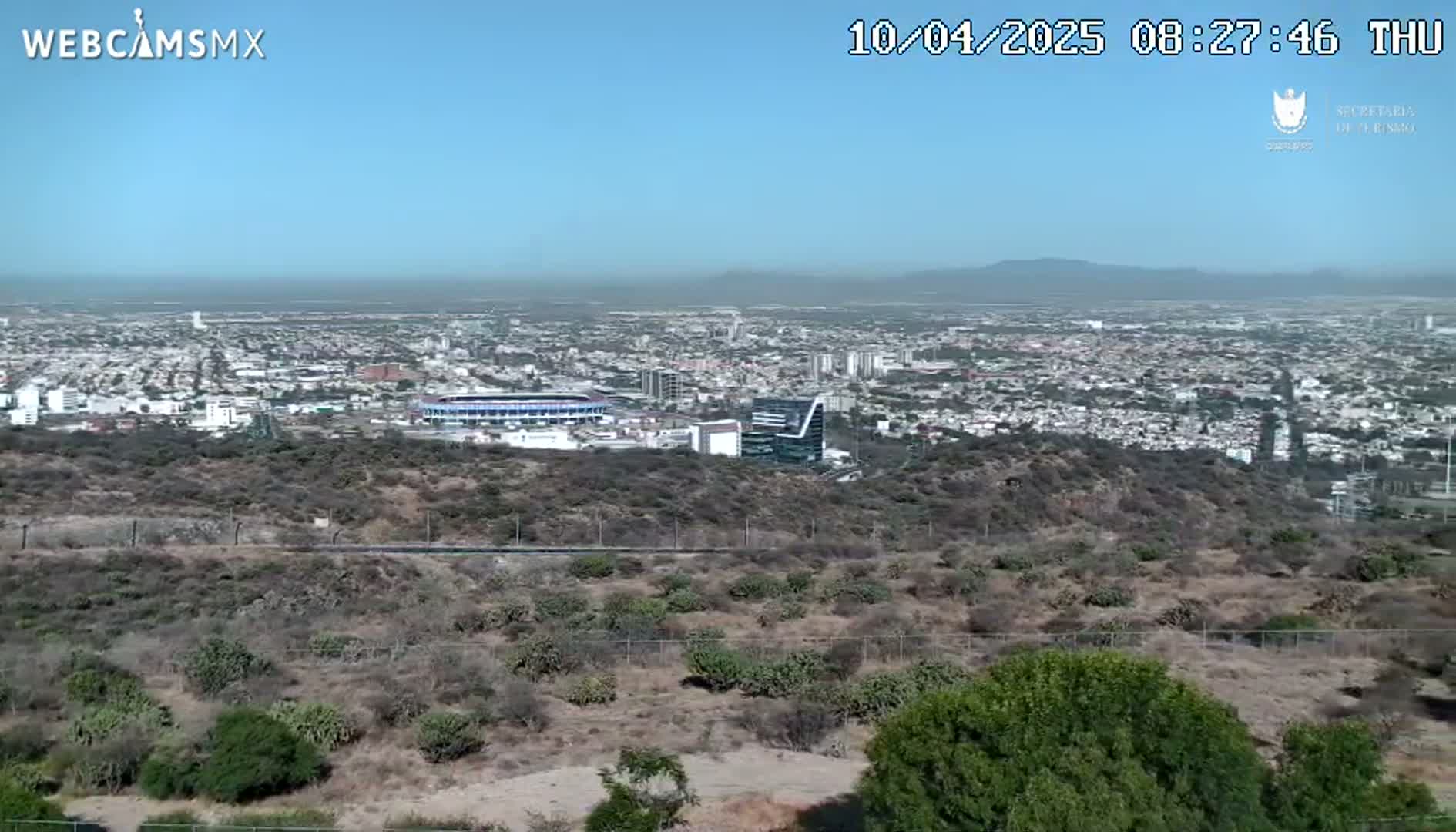 A panoramic view on a clear day shows a sprawling city with a stadium and hills in the foreground.