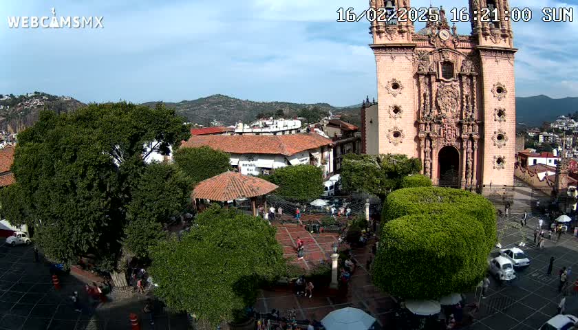 A plaza with a large church, numerous trees, and people milling about on a sunny day.