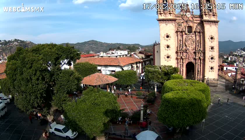 A town square with a large church, surrounded by trees and buildings, under a mostly sunny sky.