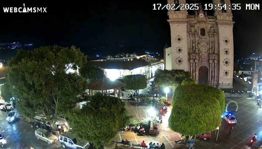 A nighttime view of a town square with a large church, surrounded by trees and people, under a clear sky.