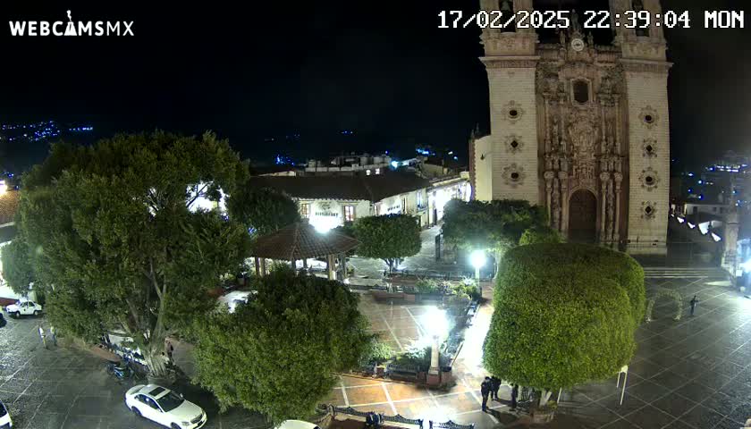 A nighttime view of a town square with a large church, numerous trees, and a gazebo, under a clear sky.