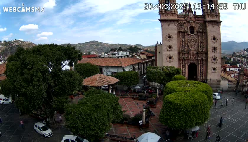 A town square with a large church, numerous trees, and several parked cars is visible under a partly cloudy sky.
