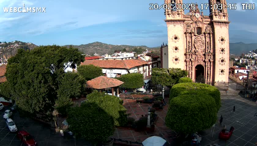 A town square with a large, ornate church, several trees, a gazebo, and cars parked nearby, under sunny skies and clear weather.