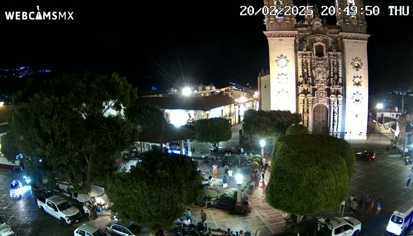 A nighttime aerial view of a town square with a large church, illuminated buildings, several parked vehicles, and people gathered in the plaza under clear skies.