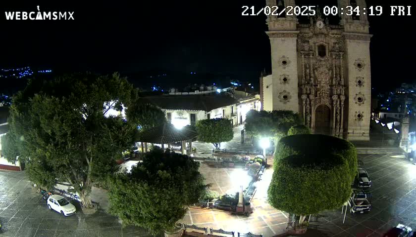 A nighttime view of a town square with a large church, several cars parked, and lush green trees under clear skies.