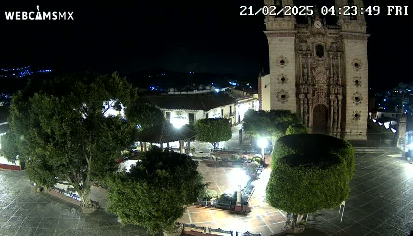 A nighttime, high-angle view shows a town square with a large church, trees, and lights, under a clear sky.