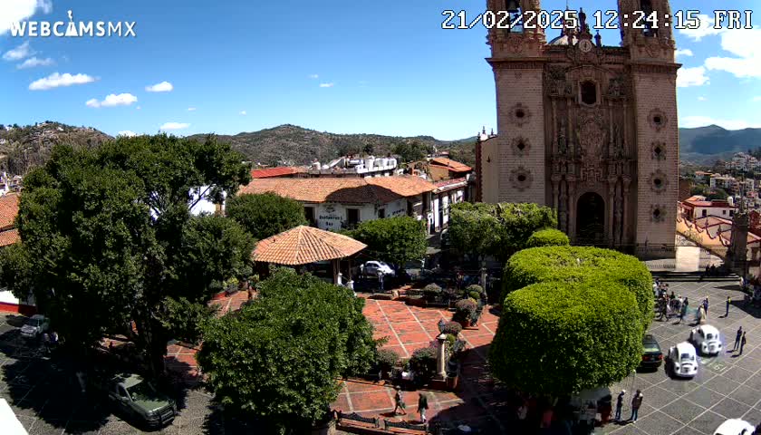 A sunny town square with a large church, several trees, a gazebo, and a few parked cars.