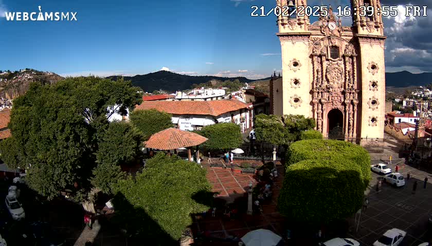 A sunny day reveals a town square with a large church, surrounded by trees and buildings, with a few people and cars visible.