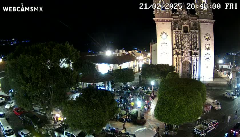A nighttime aerial view of a town square, illuminated by lights, shows a large church, trees, several parked cars, and a gathering of people.