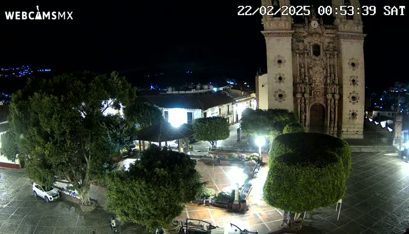 A nighttime aerial view of a town square, centered on a large church, with lush green trees and shrubs, and cars parked near the edges.