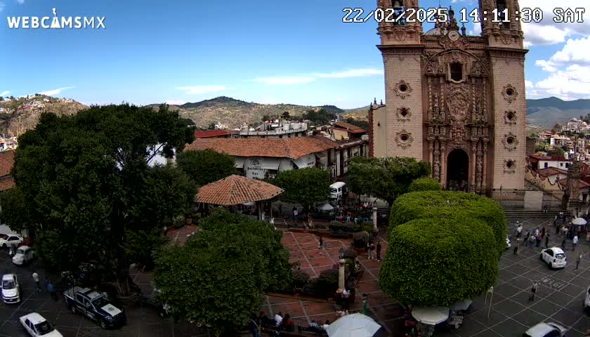 A town square with a large church, surrounded by trees and buildings, under a mostly sunny sky.
