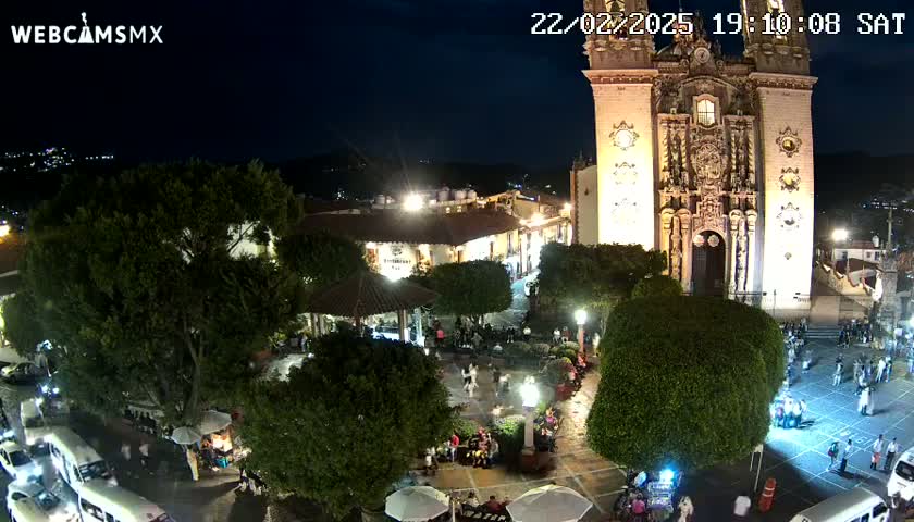 A nighttime view of a town square with a large church, illuminated buildings, and people gathered in the plaza under a clear sky.