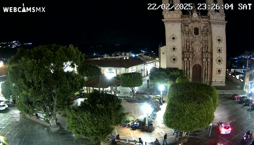A nighttime view of a town square, featuring a large church, lush greenery, a gazebo, and a few parked cars under clear skies.