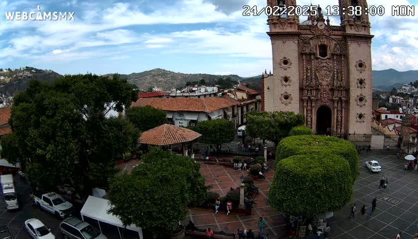 A partly sunny day shows an aerial view of a town square with a large church, several buildings, green trees, a gazebo, and people walking around.