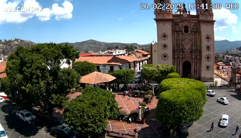 A town square with a large church, several cars, and lush green trees under a sunny sky.