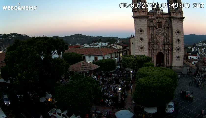 An aerial view at dusk shows a town square with a large crowd of people gathered around a gazebo near a large stone church, all surrounded by lush green trees and buildings under a clear, twilight sky.