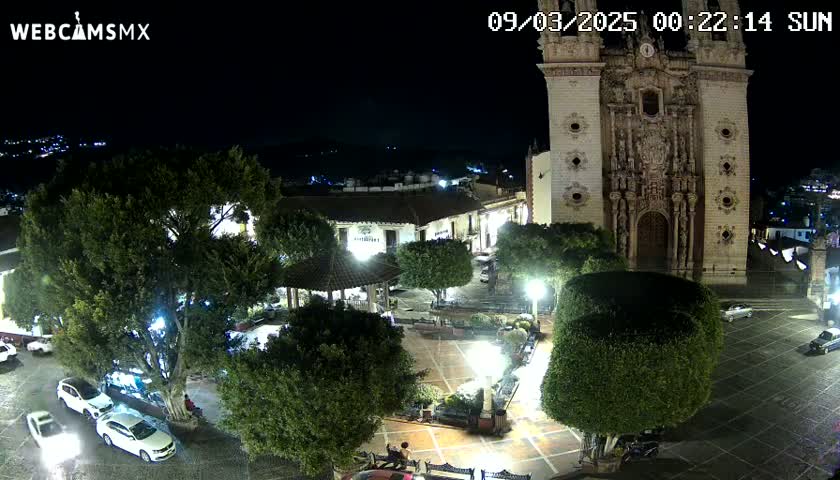 A nighttime aerial view of a town square, illuminated by streetlights, features a large church, several parked cars, and lush green trees surrounding a gazebo.